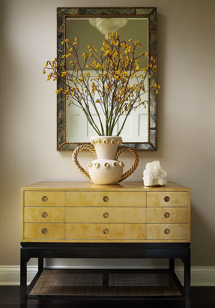 An elegant, stately, and dramatic entry way with a large sideboard or cabinet on which sits a beautiful vase with a tall natural arrangement, and on the wall is a large mirror