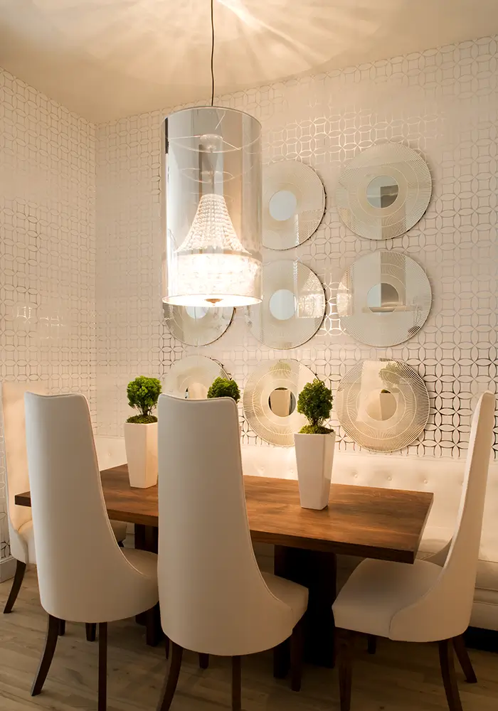 A bright, white & mirrored modern dining space with high-back white chairs, a white bench, and a series of mirrored discs on the wall, over a geometric white & silver wallpaper